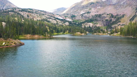 aerial drone flies over woman standing on the end of a fallen tree branch in a clear blue water lake to ravleal mountain peak and thick pine tree forest in nederland colorado rocky mountains