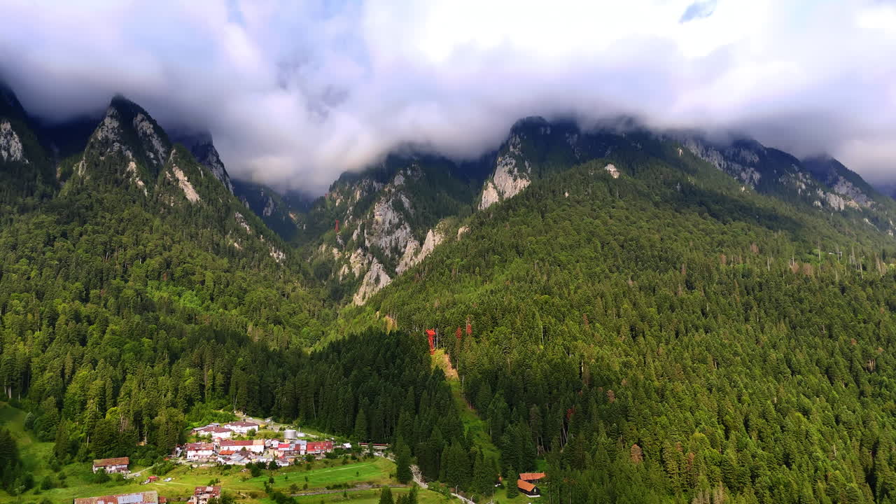 Paesaggio montano con villaggio e alberi sotto un cielo nuvoloso in Romania. Le nuvole aleggiano sulle montagne mentre un piccolo villaggio si trova tra gli alberi verdi nel paesaggio rumeno.