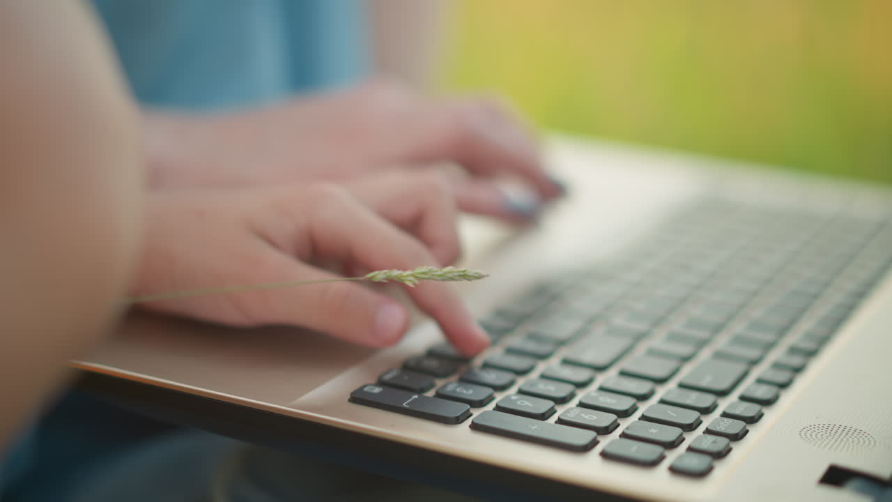 A close-up shot of a woman's hand in a blue gown, sitting on a grassy area, typing on a laptop. The image captures the moment as she gently stops a child's hand from pressing the keyboard