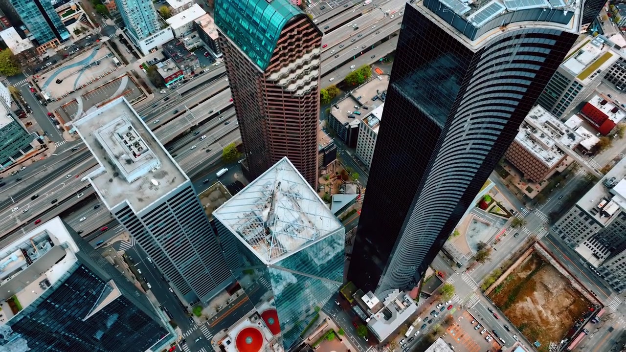 Espetaculares arranha-céus mais altos no centro de Seattle, Washington, EUA. Drone subindo sobre o topo das torres. Autoestrada de várias pistas ao fundo com tráfego circulando.