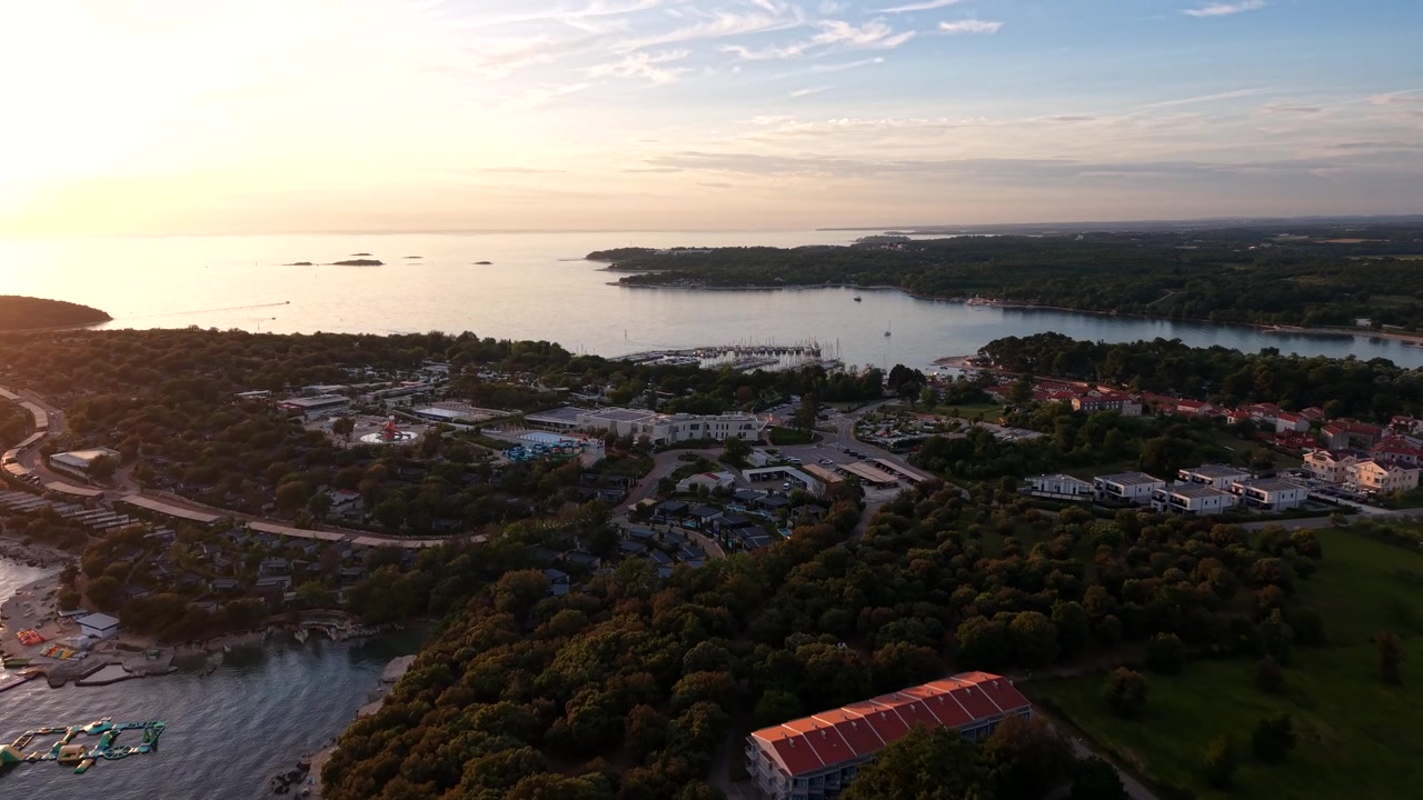 Luksusresorthotel og campingområde langs en skovklædt kystskråning nær Funtana, Istrien, Kroatien, med små øer spredt ud over det glødende Adriaterhav ved solnedgang. Bredt luftfoto med drone