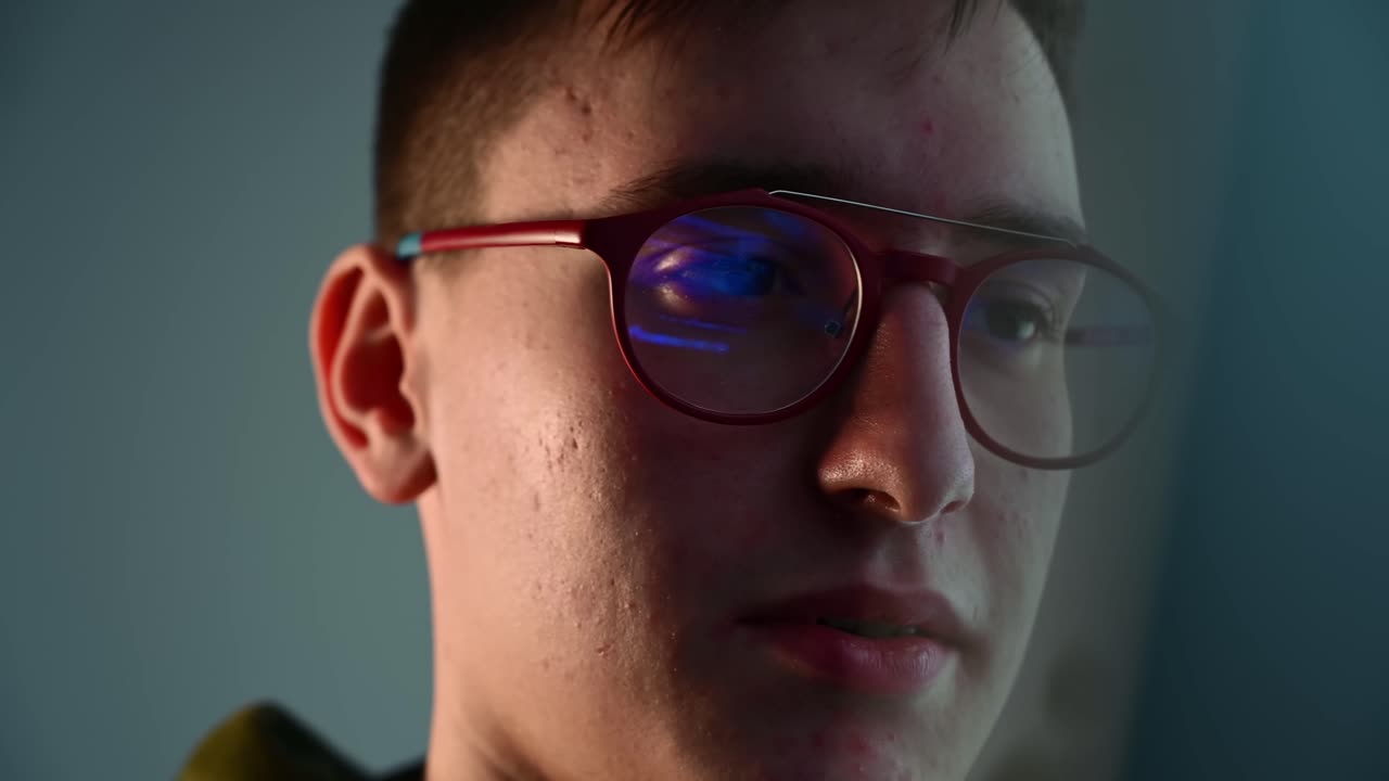 Young pimply man in red glasses watching at computer monitor in a blue lighted room. Reflection in the lens. Warm light on half of the face. Cinematic move. Close up