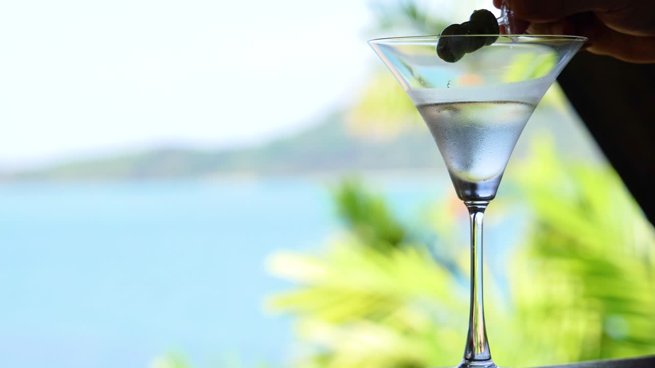 A hand places an olive in a martini glass against a sunny seaside backdrop, capturing a serene moment