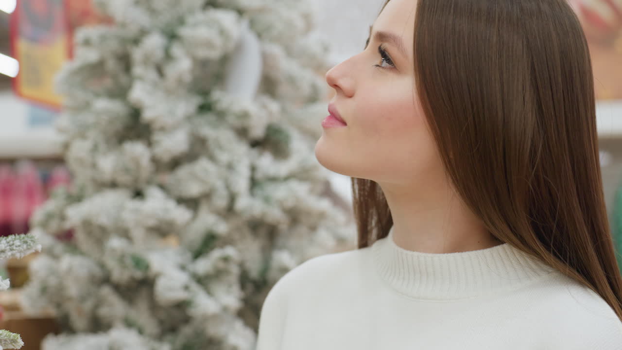 Close-up of young woman in white sweater admiring snowy Christmas tree in holiday store, she gazes at festive decorations with a peaceful expression, surrounded by a warm holiday atmosphere