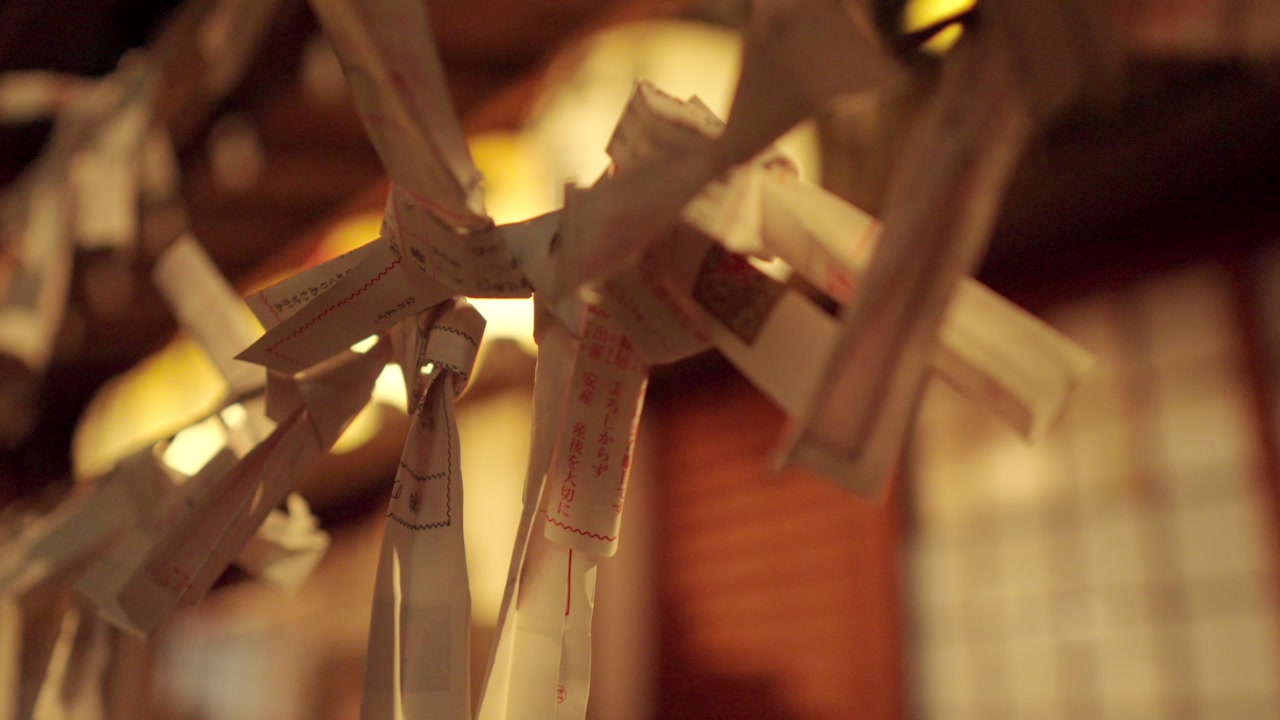 A tilt down shot of Omikuji fortune papers hanging at night at Nishiki Tenmangu Shrine in Kyoto, Japan. The papers are focused with a beautifully blurred background, capturing the mystical atmosphere