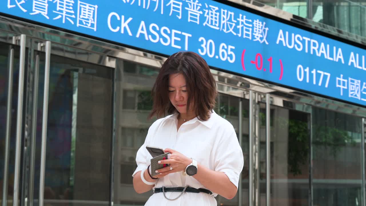 A low-angle view captures a businesswoman using a smartphone in the financial hub of Exchange Square (HKEX), home to the Hong Kong Stock Exchange, with flashing stock ticker symbols in the background.