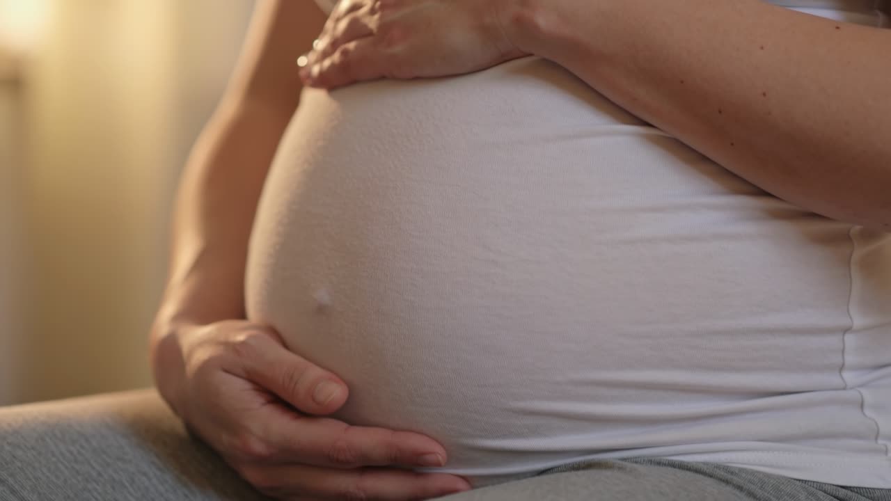 Primo piano di una donna incinta che si tiene la pancia, indossando una camicia bianca in una luce soffusa.