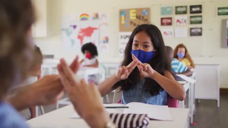 Maestra-Y-Niña-Con-Máscaras-Hablando-Entre-Sí-A-Través-Del-Lenguaje-De-Señas-En-Clase