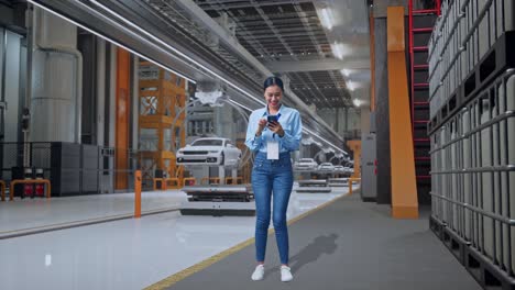 cuerpo lleno de una trabajadora profesional asiática de pie con su smartphone en la fábrica de automóviles ev, instalación industrial de alta tecnología, trabajando continuamente