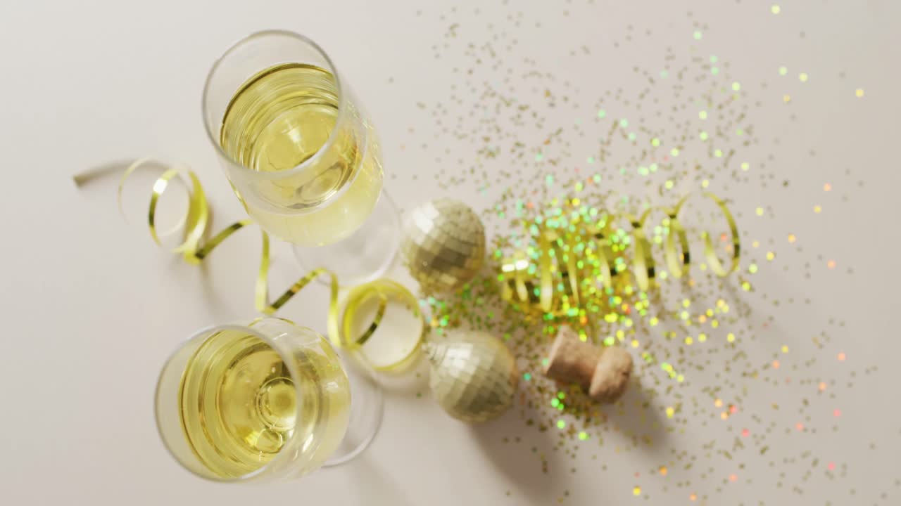 Video dall'alto di due bicchieri da champagne e tappo con stelle filanti dorate, glitter e addobbi su sfondo bianco