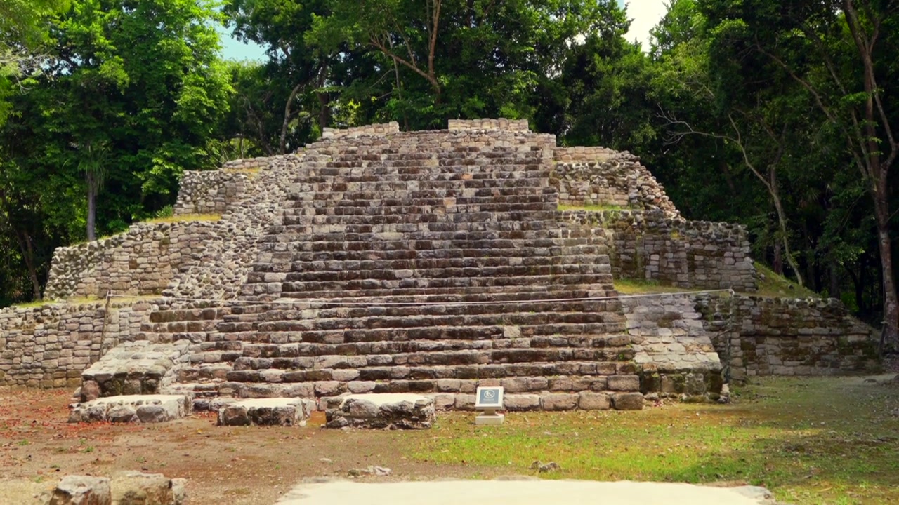 Inclinazione fluida verso il basso che rivela un'antica piramide Maya e rovine archeologiche circondate da una lussureggiante giungla tropicale nel sito di Oxtankah in Messico.