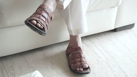 Close up of legs of an Indian man dressed in traditional white kurta pyjama and brown leather sandals, as he shakes his leg slightly, and wiggles his toes