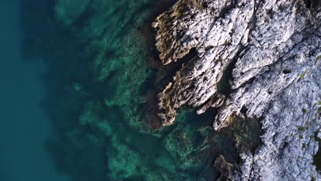 Rocky-shore-with-calm-crystal-clear-blue-water,-top-down