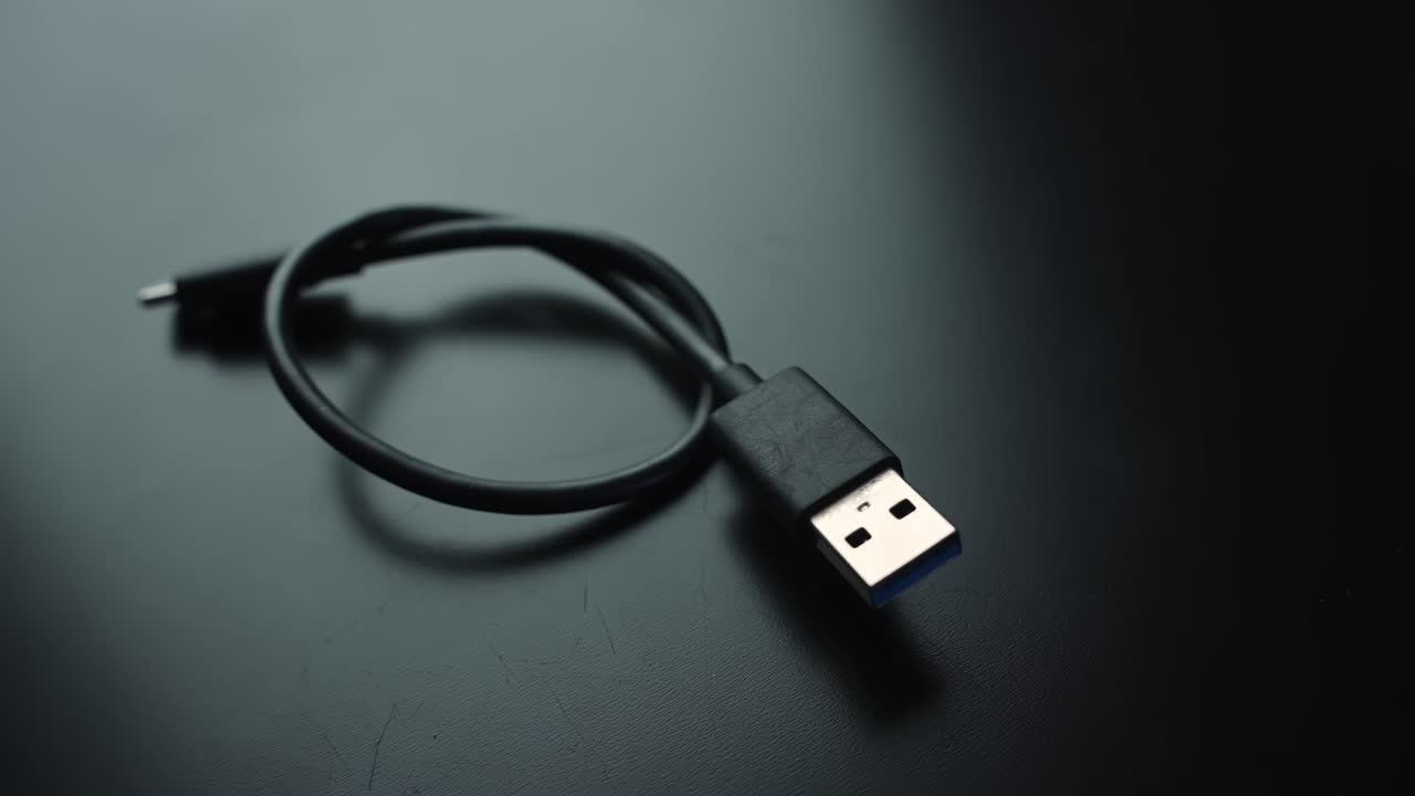 Macro close-up of a coiled black USB cable with USB-A connector on a dark desk, shallow depth of field and copy space for tech, office, and connectivity b-roll