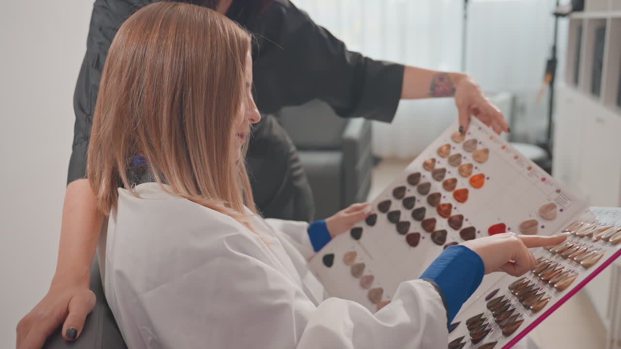 Caucasian woman choosing hair color swatches, seated in salon robe while colorist points at color chart and guides shade selection. Soft natural light fills modern interior, mood calm