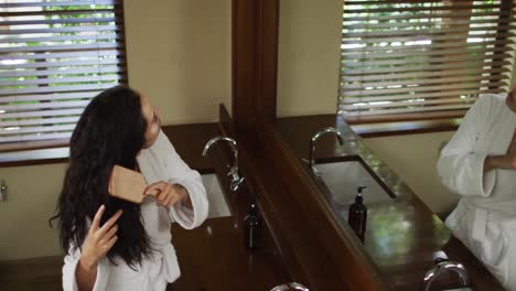 Biracial-woman-with-vitiligo-wearing-robe-looking-into-mirror-and-brushing-hair-in-bathroom