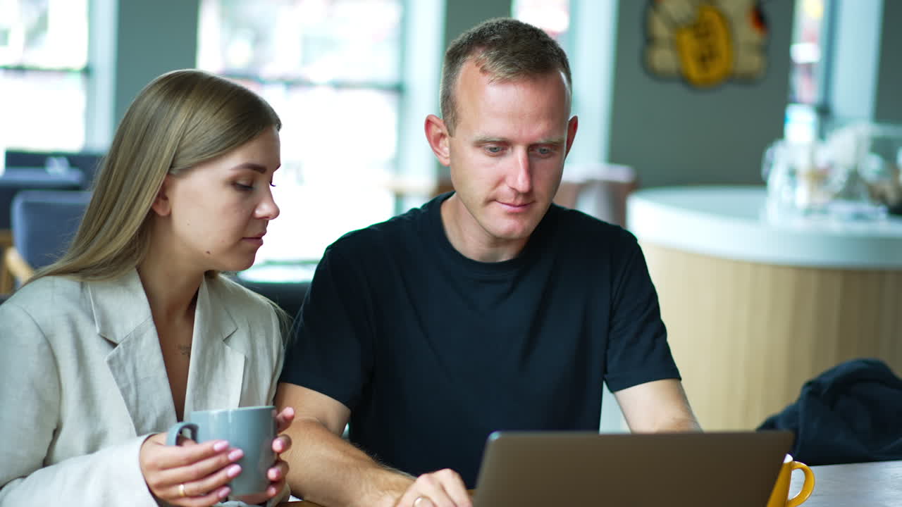 Couple sitting in a café with cups in their hands. Mid-aged young people working on laptop, discussing job. Blurred backdrop.
