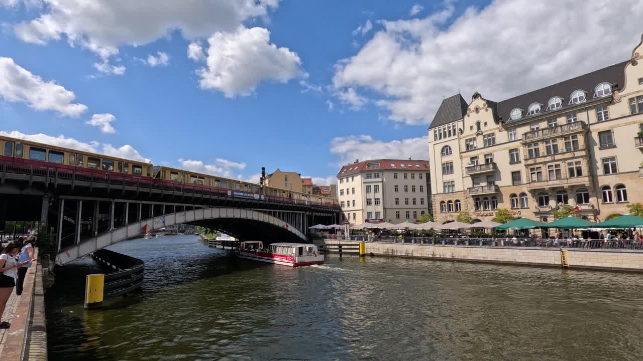 A sightseeing boat glides beneath a city bridge along a river, with pedestrians, historic buildings, and bright daylight creating a lively urban atmosphere