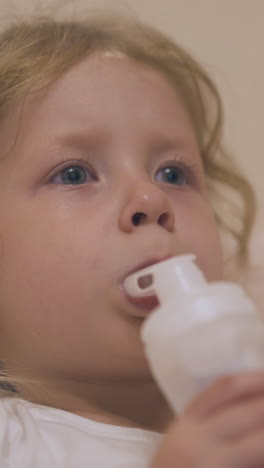cute unhappy blonde girl breathes with inhaler to cure bronchitis lying in bed on blurred background extreme close view