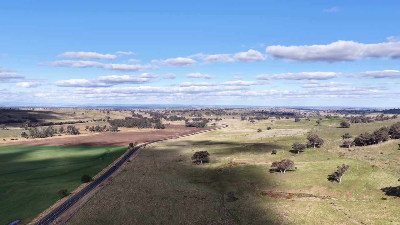 Aerial drone footage glides over open farmland and a rural road under bright daylight, revealing fields, scattered trees, and expansive blue sky