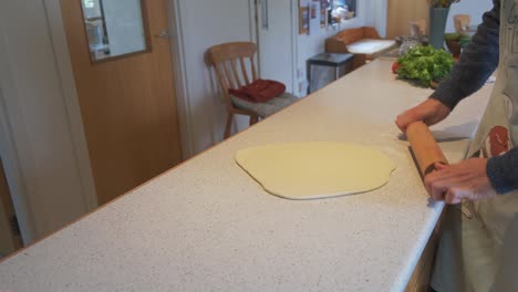 a white male cook in an apron rolls out dough with a wooden pin on his kitchen counter, flattening it into a smooth, round shape in his home kitchen.
