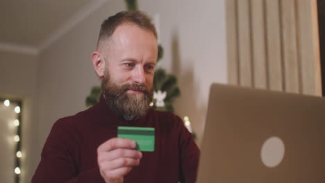 vista inferior de un hombre de cabello rojo que compra en línea con una tarjeta de crédito usando una computadora portátil sentada en una mesa en una habitación decorada con un árbol de navidad