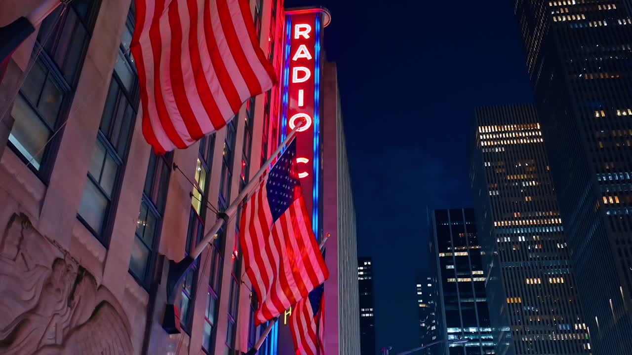 New York, USA, 12 December 2025: American flags flapping in the wind on the building facade. Radio city billboard at backdrop. Night sky over New York at backdrop