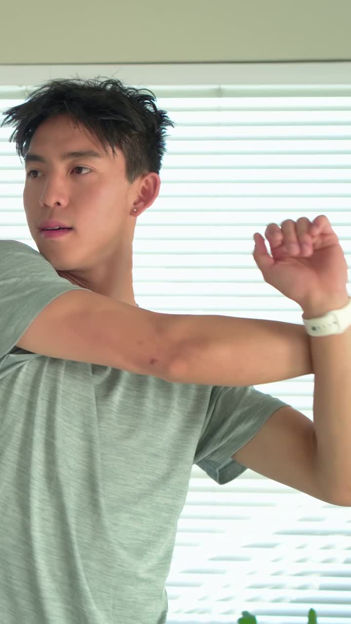วิดีโอแนวตั้ง: กำลังยืดคอ ไหล่ ข้อมือ โดยเริ่มจากการยกแขนขึ้น ชายสวมเสื้อยืดสีเทาอยู่ข้างมู่ลี่