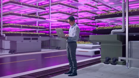 vista lateral completa del cuerpo de un trabajador profesional masculino asiático de pie con su portátil en una granja vertical, instalación industrial de alta tecnología, escribiendo en el teclado de su portátil con meditación
