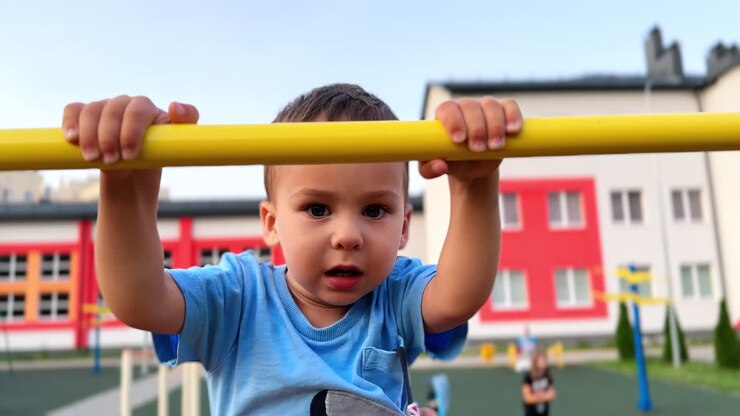 Beautiful baby boy climbs by the bars at playground. Little kid is unhappy when hold him by the waist. Close up.