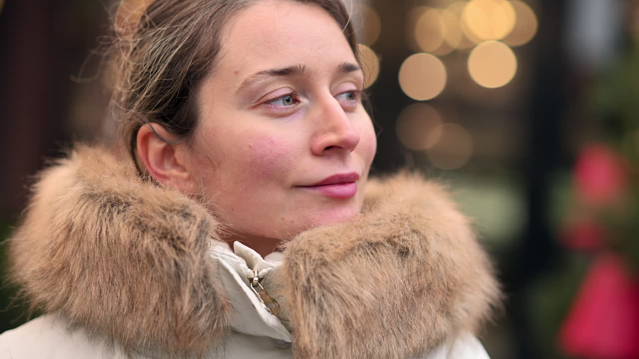 A woman stands outdoors on a chilly day, smiling while bundled in a warm coat and gloves. The background features festive decorations, creating a cheerful atmosphere