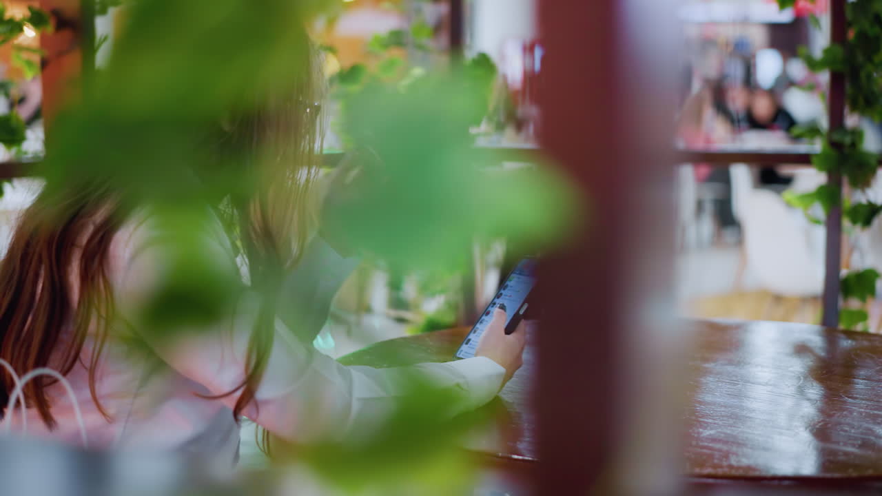 식당의 테이블에 앉아있는 여인과 함께 초록색의 클로즈업, 전화를 사용하는 동안 코를 만지고, 캐주얼하고 편안한 레스토랑 환경에서 다른 사람들을 보여주는 흐릿한 배경