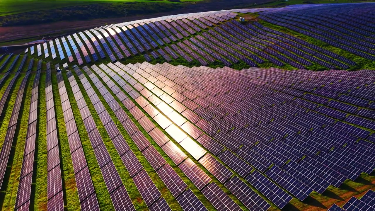 Sollys reflekteres på solpanelrækker på en bjergskråning, vist fra en diagonal luftvinkel med en stærk genskin på celleanlægget og grønt græs.