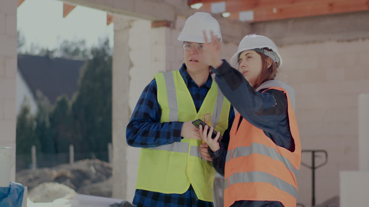 Young male and female coworkers wearing hardhats sharing smartphone while discussing at construction site