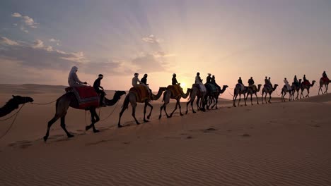 A low-angle video captures a caravan of camels with riders traversing a vast desert landscape