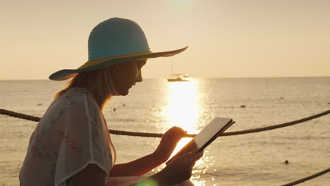 a woman uses a tablet on vacation. sits against the background of the rising sun above the sea. always in touch concept