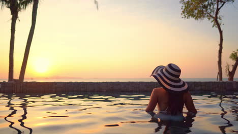 Mujer-Solitaria-Parada-En-La-Piscina-Con-Sombrero-De-Verano-Y-Disfrutando-De-La-Vista-De-La-Puesta-De-Sol-Sobre-El-Mar-Tropical