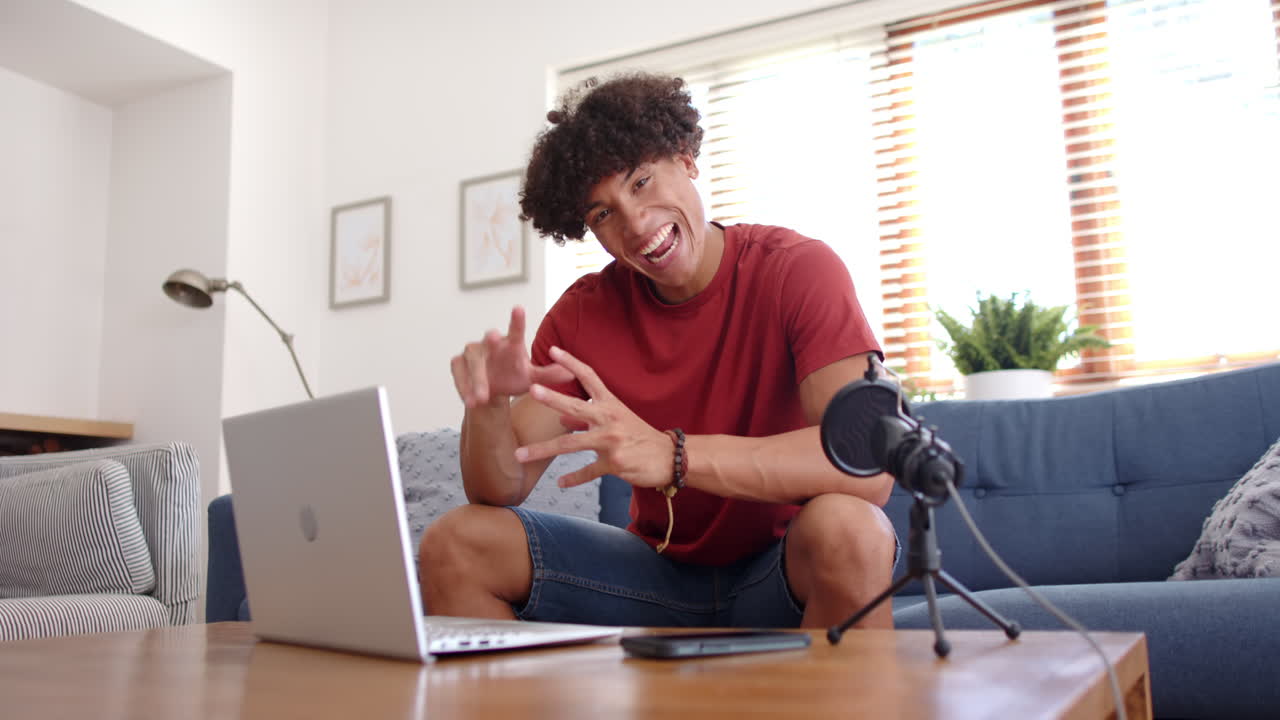 Waving and smiling, man recording podcast with microphone and laptop in living room, at home