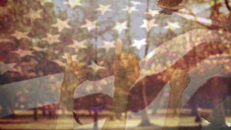 american waving flag and hands showing thumbs up over autumn leaves falling against park