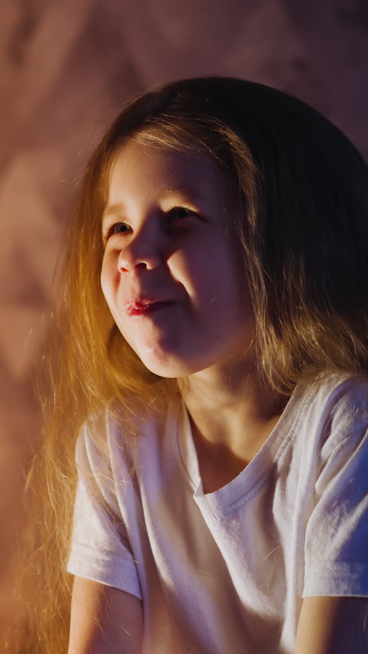 Pretty little girl with long loose hair chews food looking at parent while spending time together in semidark room with glowing light lamp slow motion
