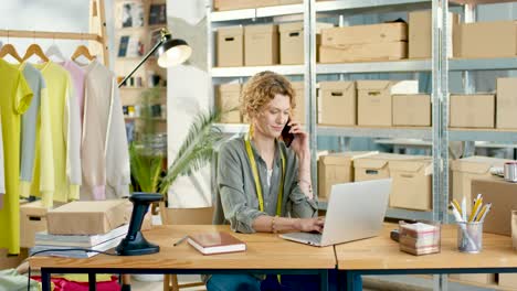 close-up view of caucasian designer woman sitting at desk talking on smartphone and working on laptop in clothing shop warehouse