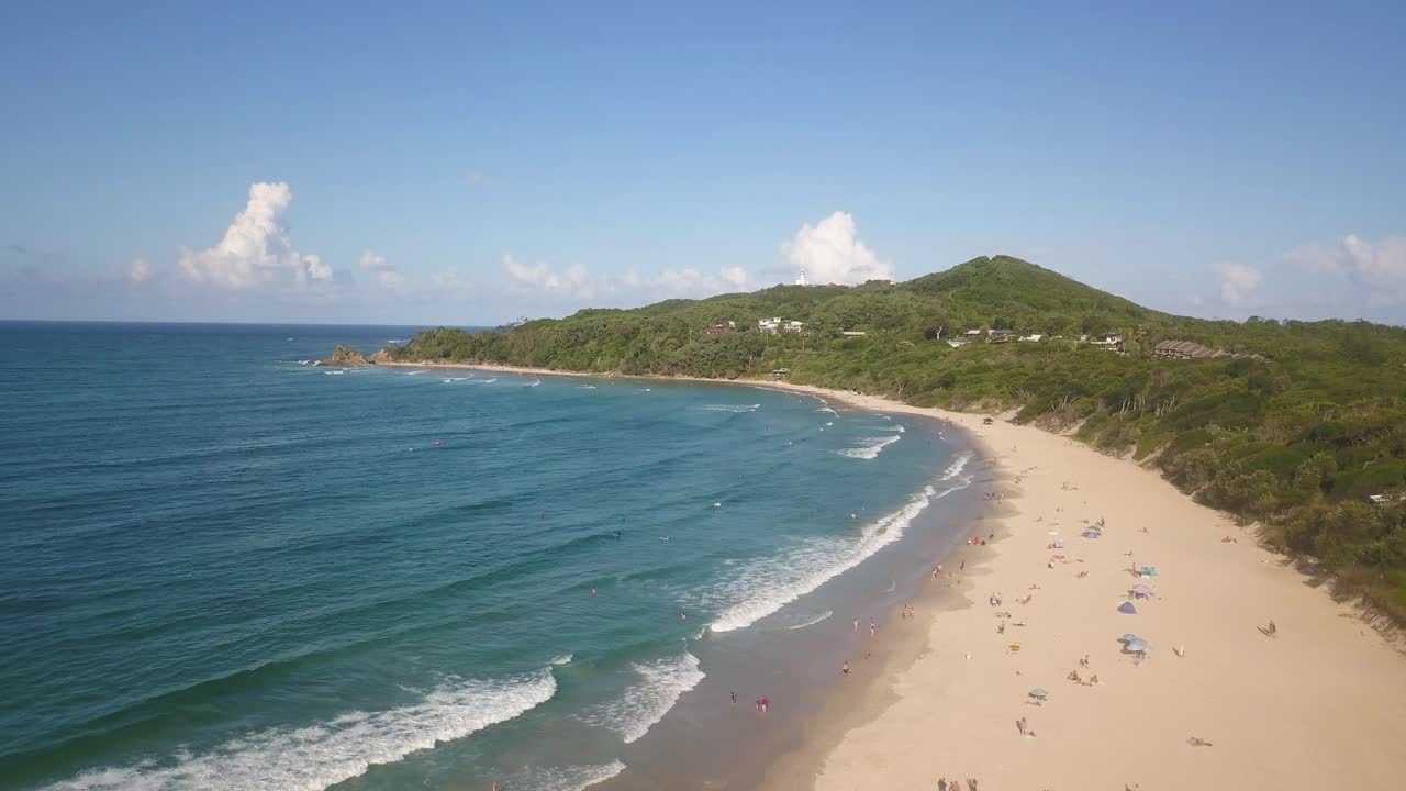 은 날에 바이런 베이 해변으로 내려가는 공중 파노라마,  모래 해변, 색 물, 그리고 관광객들이 육지에서 휴식을 취하는 자연 환경을 보여줍니다.