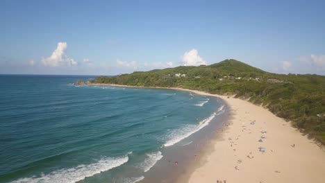 panorámica aérea desciende a la playa de la bahía de byron en un día soleado, mostrando su playa de arena blanca, aguas turquesas, y el entorno natural como los turistas se relajan en la tierra