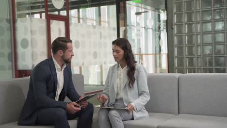 a young businessman with beard and suit has a business meeting with a pretty young businesswoman on the sofas in the common area of the office building