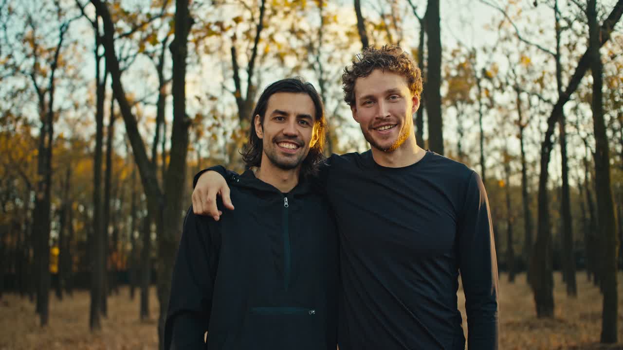 Portrait of two happy guys with black hair and beard in black sportswear who are on a morning jog in the autumn forest in the morning. Two guys are hugging and posing in the morning In the forest