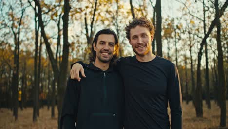 Portrait-of-two-happy-guys-with-black-hair-and-beard-in-black-sportswear-who-are-on-a-morning-jog-in-the-autumn-forest-in-the-morning.-Two-guys-are-hugging-and-posing-in-the-morning-In-the-forest