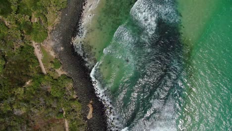 Mirando-Verticalmente-A-Las-Pequeñas-Olas-Rompiendo-En-La-Playa-Rocosa-Adyacente-A-La-Pista-Para-Caminar-Brillante-Día-De-Verano,-Drone