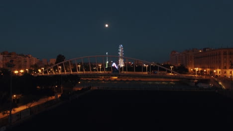 Vista-Aérea-Nocturna-De-La-Noria-Iluminada-Y-El-Puente-Contra-El-Cielo-Con-Luna-Valencia-España