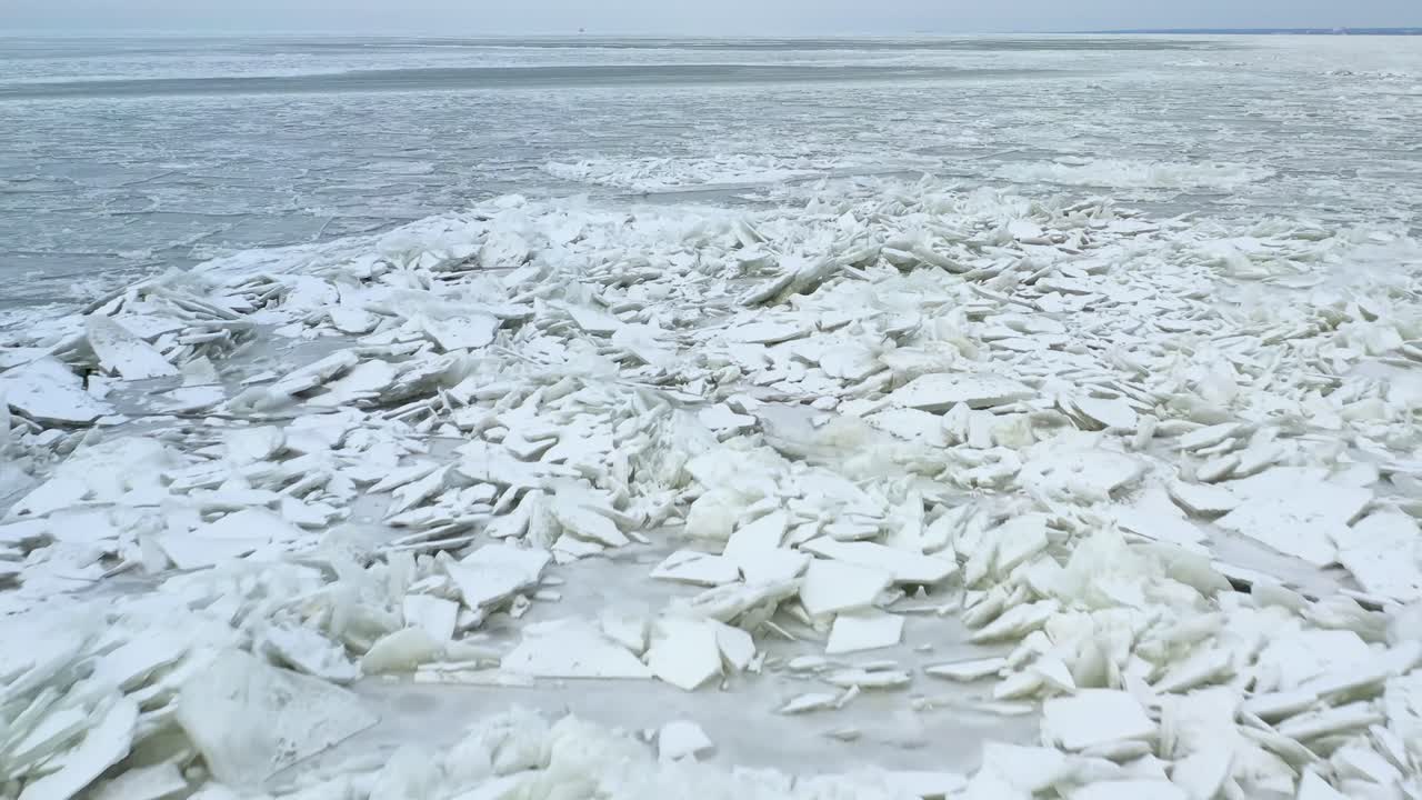 Drone flying backwards over sheet of melting ice on lake. Miniture iceburgs melting on water with light house far in background.