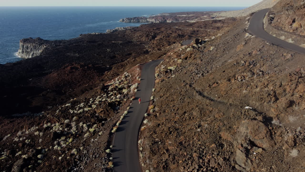 Woman walking up a mountainous hill on the road at sunset, overlooking the bay and blue horizon, Hierro Island, Canary - view from above